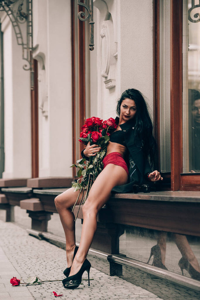 Elegant beautiful brunette woman in red shorts and black coat girl posing sitting on the windowsill with a big bouquet of red roses. Birthday. Valentines day. Selective focus, filmgrain - Foto, imagen