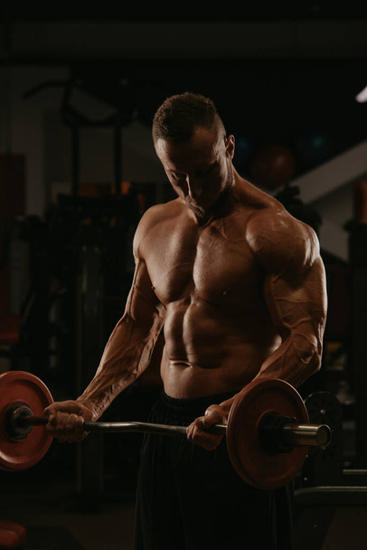 Um fisiculturista com barba está fazendo cachos de bíceps com uma barra curta em um ginásio. Uma foto de um tronco de um homem musculoso nu durante um treino. - Foto, Imagem