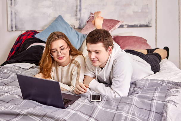 Online shopping using laptop for young couple lying on their stomachs in bed in their bedroom. - Foto, Imagem