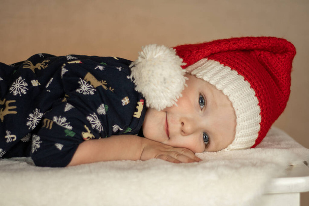 a little cute boy in a santa hat and New Years pajamas lies on a fur rug on a plain background - 写真・画像
