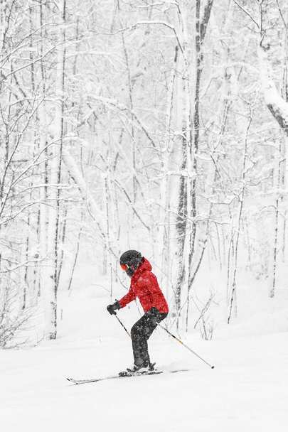 Alpine skiing. Woman skier going dowhill fast against snow covered trees background during winter snowstorm. Woman in red jacket and goggles. Winter wonderland - Фото, изображение