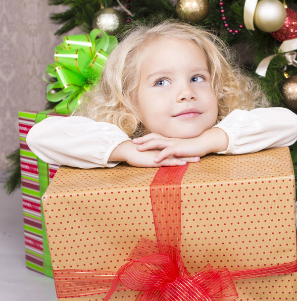 cute little girl holding a gift box and sitting beside a Christmas tree - Fotó, kép