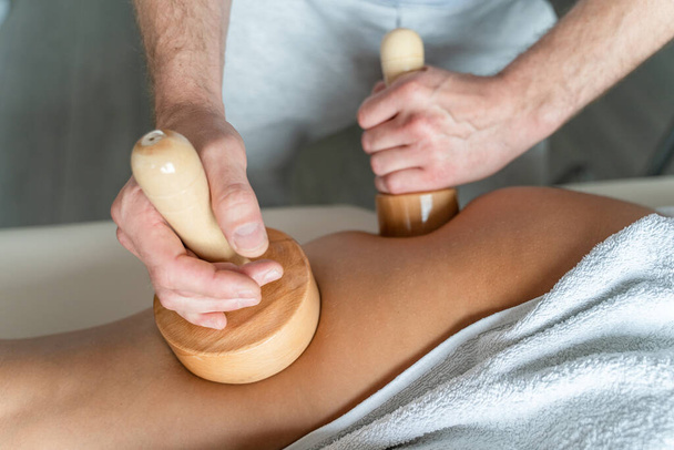 Close up on back of the leg of unknown woman having madero therapy massage anti-cellulite treatment by professional therapist holding wooden tools in hands in studio or salon with copy space - Fotó, kép