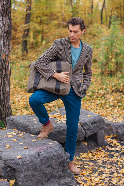 Tall handsome man dressed in a brown jacket posing with a bag near the rocks in autumn forest - Foto, immagini