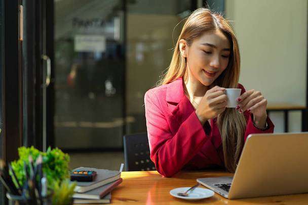 Vonzó ázsiai fiatal üzletasszony pihen a munkahelyén, forró teát vagy kávét kortyolgat miközben a laptop képernyőjét nézi. - Fotó, kép