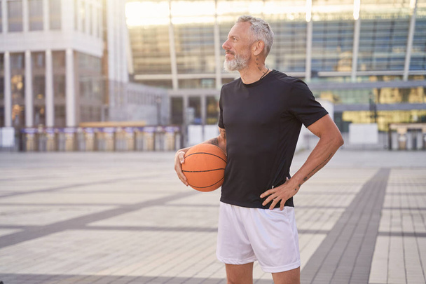 Przemyślany sportowy dojrzały mężczyzna w odzieży sportowej stojący na zewnątrz z piłką do koszykówki, gotowy do treningu - Zdjęcie, obraz