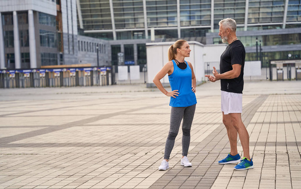 Полнометражный снимок двух спортивных друзей, мужчины и женщины среднего возраста в спортивной одежде, разговаривающих на улице, занимающихся спортом вместе в городе - Фото, изображение
