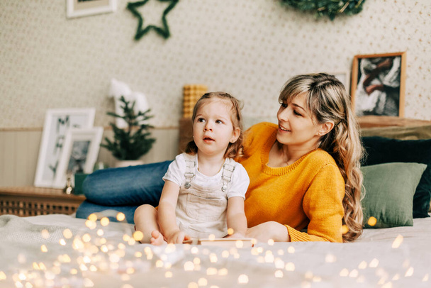 Mom and daughter are reading a book and smiling, sitting at home on the bed. New Year, Christmas, family, time together - Foto, Imagen