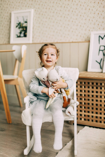 Urocza dziewczynka siedzi na krześle i przytula swoje ulubione zabawki. Dzieciństwo, zabawki, opieka - Zdjęcie, obraz