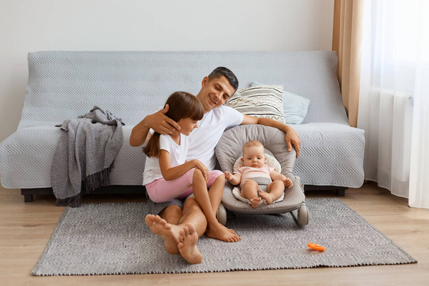 Indoor shot of happy father sitting on floor near cough with his daughters, hugging his elder kid, spending time with infant baby girl in rocking chair. - Valokuva, kuva