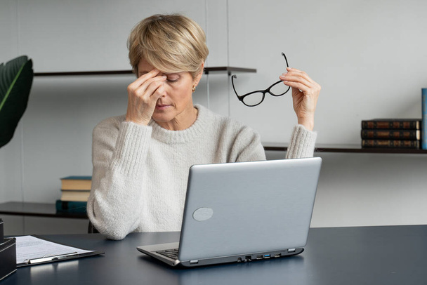 A middle-aged business woman, sitting at her desk, takes off her glasses from the pain in her eyes and massages the bridge of her nose. - Photo, Image
