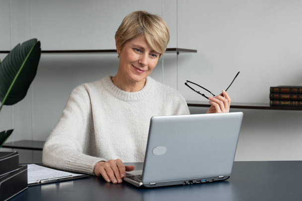Portrait of a mature businesswoman working at a laptop in the workplace in the office, takes off her glasses and thinks while looking at the laptop - Fotografie, Obrázek