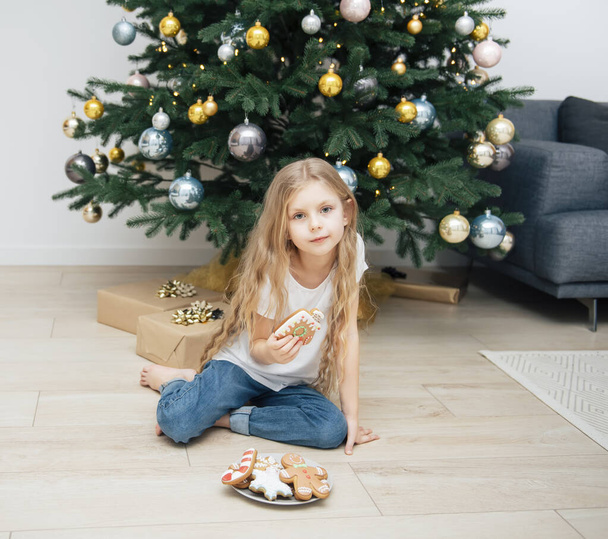 Girl eating Christmas gingerbread near the Christmas tree in the living room - 写真・画像
