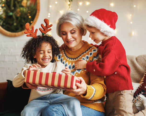 Farklı ırklardan iki çocuk Noel ağacının yanında otururken büyükannesine yeni yıl hediyesi veriyor. - Fotoğraf, Görsel