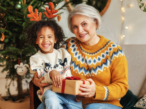 Mutlu Afro-Amerikan kıvırcık çocuğu büyükannesinin kucağında oturuyor, noel hediyesiyle, kameraya gülümsüyor. - Fotoğraf, Görsel