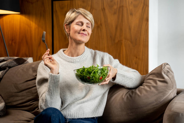 Middle-aged woman is sitting and resting at home on the couch, eating a green salad. A woman eating healthy food. - Φωτογραφία, εικόνα