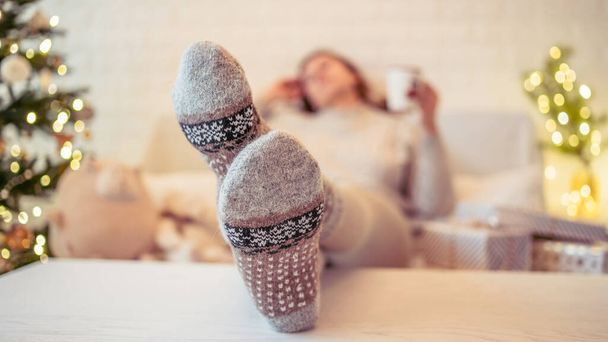 Woman sitting on couch near christmas tree. Female legs, feet in winter woolen socks putting on the table and relaxing after packing handmade  gift boxes. Presents for family. - Foto, imagen