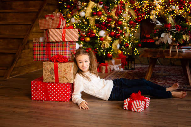 Portrait of a young girl sitting at a Christmas tree with a large pile of packed gift boxes, happy to receive many gifts on Boxing day. A mountain of gifts falls with the child. Step 5 - Photo, Image