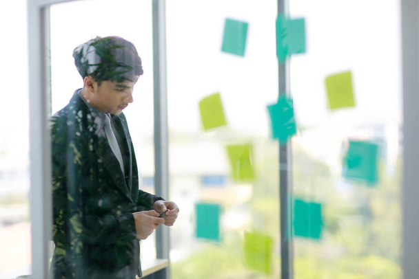 lähikuva komea aasialainen liikemies mustassa puvussa kirjoittaa ideansa tulevasta projektista tahmealla huomautuksella vakavalla tuulella modernissa toimistossa.. - Valokuva, kuva