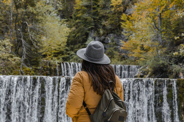 A Caucasian woman with autumn clothes and backpack standing in front of the waterfall - 写真・画像