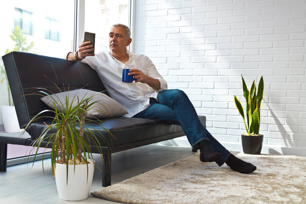 Satisfied man relaxing in his living room using mobile phone while having coffee - Fotó, kép