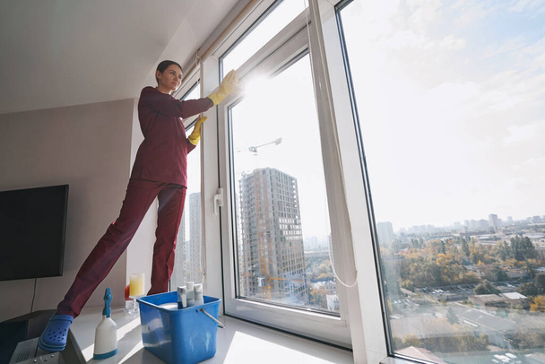 Pensive housecleaner in rubber gloves wiping window-frame - Foto, Imagen