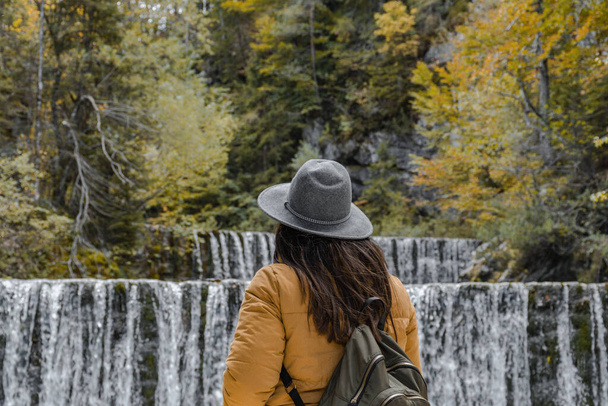 A Caucasian woman with autumn clothes and backpack standing in front of the waterfall - Foto, Imagen