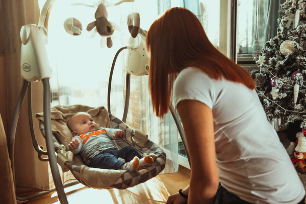 A young mother playing with her newborn kid lying in a small bed in a sunny room - Foto, imagen