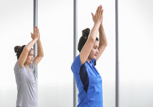 Gesunde und starke erwachsene Frauen üben Yoga-Aktivität auf der Fitnessmatte, indem sie Baumhaltung üben, um Körperbalance und Kraft mit einem Bein zu trainieren, das mit den Händen über dem Kopf in einer Sporthalle steht.. - Foto, Bild