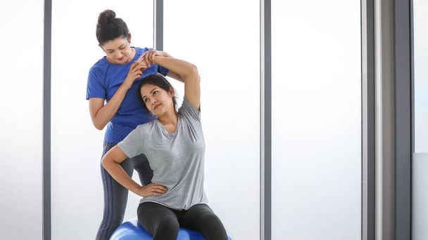 Entraîneur de yoga professionnel de charme coaching débutant féminin pour l'exercice des hanches et des épaules en aidant la femme asiatique pour la flexion des bras, pose du corps tout en étant assis sur la balle de fitness pour l'entraînement de force dans la salle de gym. - Photo, image