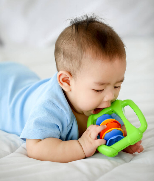 Αξιολάτρευτο και αθώο μικρό ασιατικό μωρό απολαμβάνει να παίζει διασκέδαση με πολύχρωμα παιδιά παιχνίδι, ενώ βρίσκεται στο στομάχι στο κρεβάτι. Υπέροχο νήπιο μαθαίνουν φυσικό σχήμα και απόχρωση του χρώματος αρπάζοντας αντικείμενα στο νηπιαγωγείο. - Φωτογραφία, εικόνα