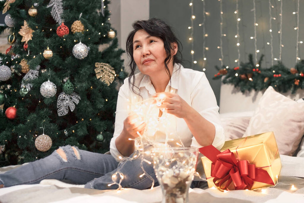 Senior asian woman on bed with xmas lights, portrait of attractive elderly lady with garland in hands and gold gift at home at christmas holiday - Foto, imagen