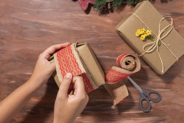 A top view of a person wrapping a Christmas present with jute fabric ribbon with red lace on a table - Foto, Imagem
