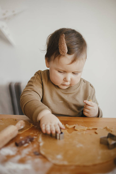 Urocza córeczka robiąca razem świąteczne pierniki na niechlujnym drewnianym stole. Cute dziewczynka maluch pomaga mamusi cięcia ciasta z świąteczne frezy do ciasteczek. Czas rodzinny - Zdjęcie, obraz