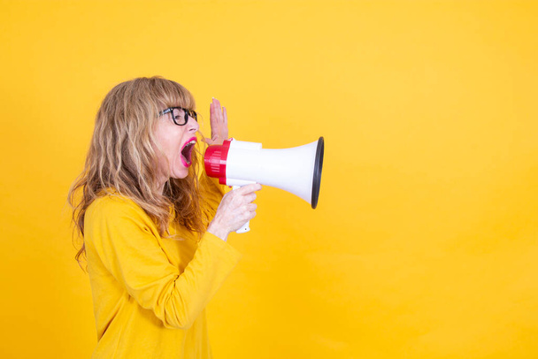 mature woman yelling with megaphone with yellow background - Valokuva, kuva
