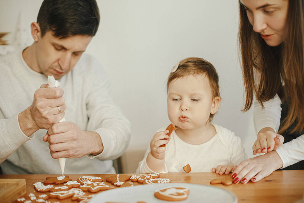 Happy stylish family decorating christmas gingerbread cookies with icing on wooden table. Cute little daughter, mom and dad time together. Xmas holiday preparations. Authentic lovely moments - Photo, Image