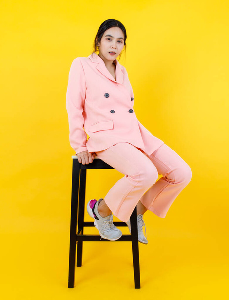 Pembe pastel ceketli ve pantolonlu yaramaz Asyalı kız yetişkin kostüm modası için yüksek sandalyeye rahatça oturur. Modern kıyafetler için rahatlatıcı bir portre ve kadın kıyafeti.. - Fotoğraf, Görsel