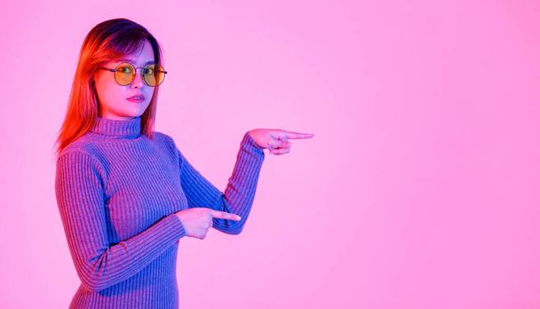 Studio ampui Aasian nuori nainen malli harmaa poolopaita ja oranssi linssi aurinkolasit seisoo osoittava etusormi tyhjään kopiotilaan esittää edistää tuotteen vaaleanpunainen valo tausta. - Valokuva, kuva