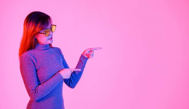 Studio shot Aziatische jonge vrouw model in grijze coltrui en oranje lens zonnebril staande wijzende index vingers om lege kopieerruimte presenteren bevorderen product op roze licht achtergrond. - Foto, afbeelding