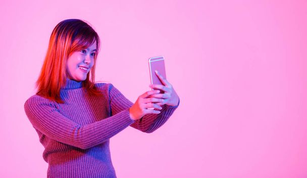 Portret studio shot van Aziatische stedelijke vrouw model in grijze coltrui staande glimlachende blik op camera met touchscreen smartphone in handen chatten met vrienden op roze licht achtergrond. - Foto, afbeelding