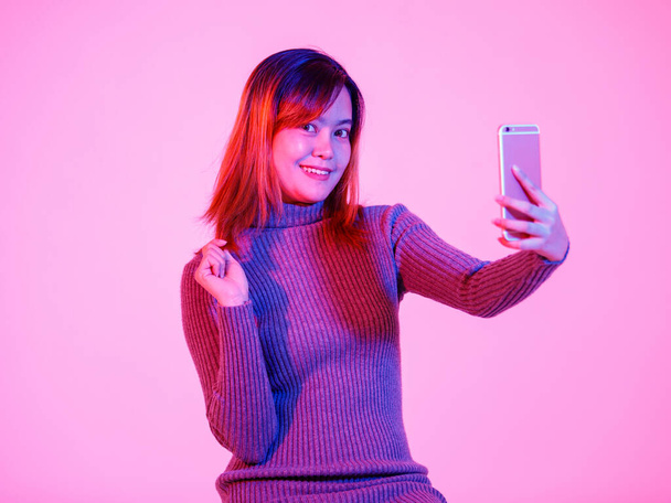 Studio shot de modelo femenino urbano asiático de moda en suéter de cuello alto gris de pie sonriendo con un teléfono inteligente sobre fondo rosa claro. - Foto, imagen