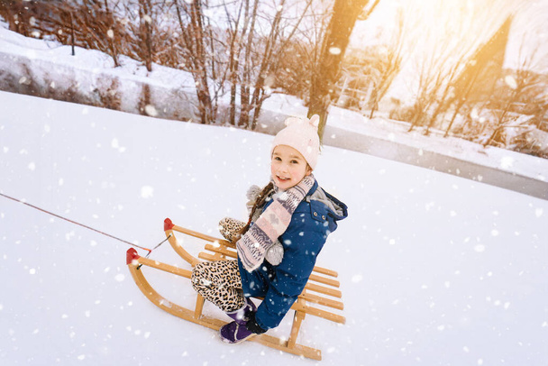 Dvě děti jezdí na dřevěných retro saních za slunečného zimního dne. Aktivní zimní hry venku. Zimní aktivity pro děti. Děti si hrají se sněhem v parku.  - Fotografie, Obrázek