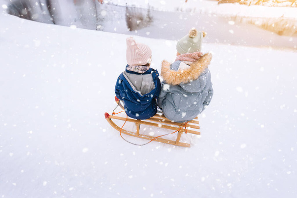 Двое детей едут на деревянных ретро санях в солнечный зимний день. Активная зима на открытом воздухе игры. Зимние мероприятия для детей. Дети играют со снегом в парке.  - Фото, изображение