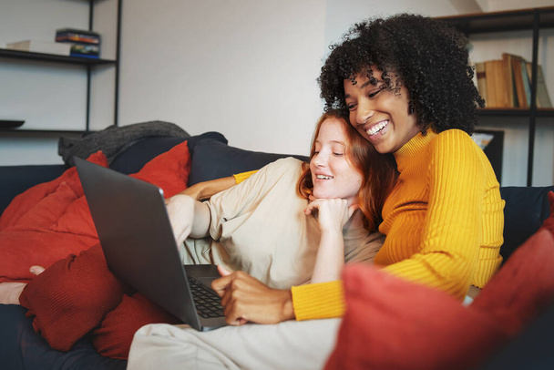 Couple of young homosexual women smiling watching content on a laptop sitting on a couch - multiracial lesbian couple in love concept - 写真・画像