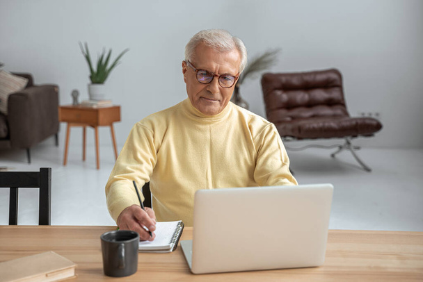 Senior man wearing glasses sitting in front of the laptop and writing notes - Valokuva, kuva