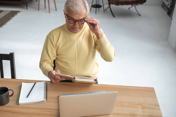 Mature man straightens glasses while reading interesting book with serious expression - Foto, immagini