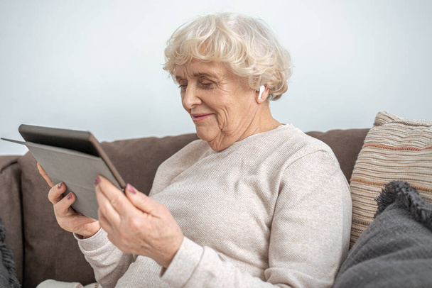 Grandmother wearing wireless earphones sitting on couch in living room and using tablet - Fotó, kép