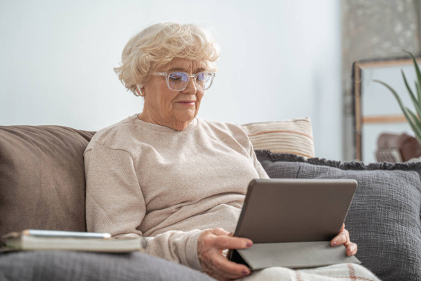 Senior woman wearing glasses sitting at the couch at home and using tablet - Foto, Imagen