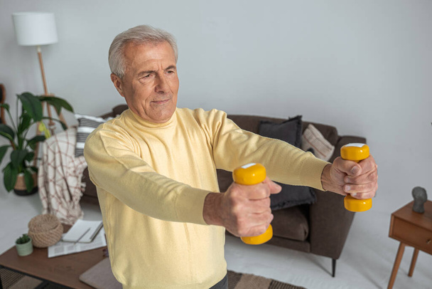 Senior man training with dumbbells at home while looking away and feeling happy - Foto, immagini