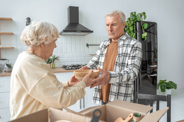 Mature man giving plate to his senior wife while unpacking boxes at the kitchen - Photo, image